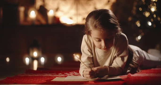 Cute little girl is lying on carpet in room decorated for christmas, writing a letter for santa, preparing for holiday - christmas spirit concept close up 4k footage