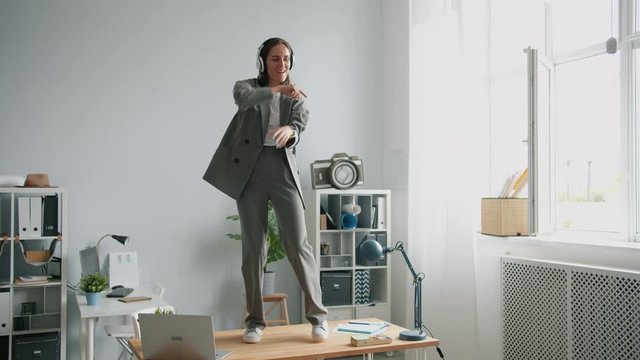 Excited young businesswoman is dancing on table in office having fun wearing headphones enjoying music relaxing at work. People, job and entertainment concept.