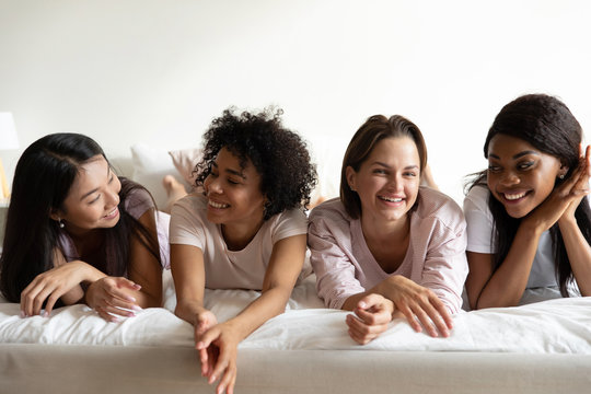 Happy diverse young ladies relax on bed enjoy pajama party