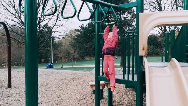 Young little Caucasian girl is scared to hang climb on monkey bar in park. Sport challenge for kid. Active preschool child doing exercise outdoor on playground. Healthy happy childhood.