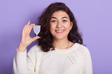 Close up portrait of young smiling beautiful woman holding menstrual cup in one hand, being in good mood, looking directly at camera with charming smile, isolated on purple. Gynecology concept.