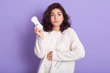 Gloomy Caucasian woman cares about personal hygiene, holds sanitary pad in hands, suffers from pain during menstruation, has sad expression, wear white outfit, poses in studio isolated over lilac wall © sementsova321