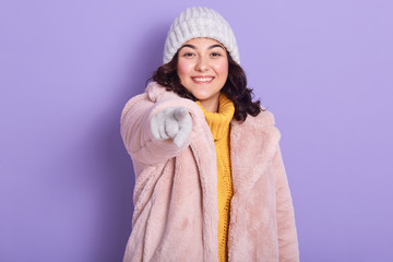 Portrait of good looking positive model reaching out her hand, showing direction, looking directly at camera, wearing yellow sweater, light hat and faux fur coat. People and emotions concept.