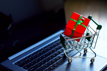 Gift box wrapped in red paper with green ribbon in small trolley, mini pushcart, cart are on laptop computer. Online shopping concept. Black friday sale. New year, christmas fair, market.