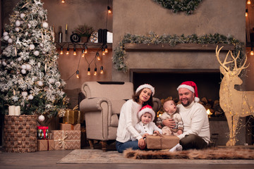 Photo of parents in Santa's cap with two sons sitting on floor by fireplace next to deer with garland in room