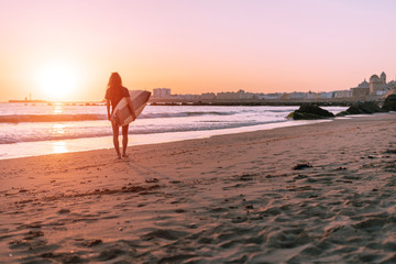 a girl going into the distance on the sand near the water carries a surf in her hand at sunset towards the sun and the city in the distance