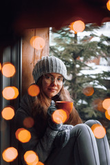 Beautiful  woman holding and drinking a cup of coffee or cocoa in gloves sitting home by the window. Blurred winter snow tree background. Morning, coziness, winter and people concept