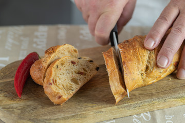 A man cuts fresh bread baguette with bran and pepperoni with a ceramic knife on a mango wood chopping board in the kitchen. A pod of pepper nearby.