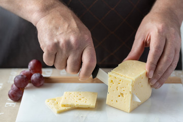 A man in a dark apron cuts a piece of genuine cheese with a ceramic knife on a marble board in the kitchen. A bunch of grapes nearby.