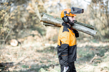 Professional lumberman in protective workwear with a chainsaw carrying wooden logs during the work...