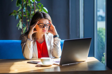 Headache, migraine, confusion, problem or thinking. Portrait of sick brunette young woman in glasses sitting, holding her painful head down and feeling bad. indoor studio shot, cafe, office background