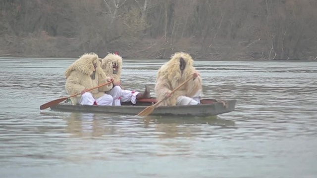 Three men in traditional hungarian buso costume calmly paddling in a boat over the Danube river in the winter time to the annual mohacs folk festival in slow motion.