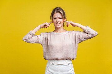 I don't want to hear you. Portrait of annoyed young woman with fair hair in casual blouse standing, closing ears with fingers and looking at camera. indoor studio shot isolated on yellow background