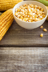 Raw corn on the cob and grains of corn in white bowl on wooden table