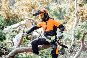 Portrait of a professional lumberman in protective workwear sitting with a chainsaw on the felled...