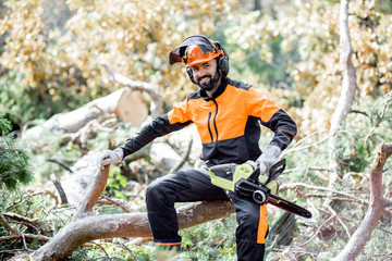 Portrait of a professional lumberman in protective workwear sitting with a chainsaw on the felled tree, resting after the hard work in the forest