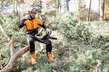 Portrait of a professional lumberman in protective workwear sitting with a chainsaw on the felled tree, resting after the hard work in the forest