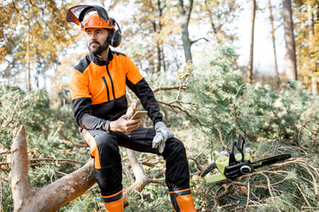 Portrait of a professional lumberman in protective workwear sitting with mobile phone on the felled...