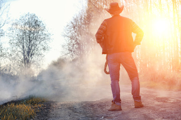 Fototapeta premium A man is wearing a cowboy hat and a loso in the field. American farmer in a field wearing a jeans hat and with a lasso in the smoke of a fire. A man walks through a burning field