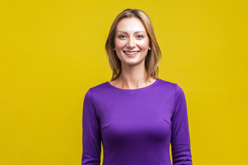 Fototapeta premium Portrait of positive elegant woman in tight purple dress standing, looking at camera with cute engaging toothy smile, beautiful with clean shiny skin. indoor studio shot isolated on yellow background