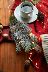 Obraz na płótnie Canvas A cup of coffee and a garland on a wooden tray with a cozy red sweater and a warm hat. New Year's still life with a fir branch and mountain ash