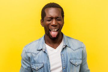 Portrait of positive carefree young man in denim casual shirt standing with closed eyes and showing out his tongue, foolishing and making face. indoor studio shot isolated on yellow background