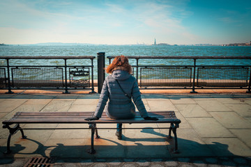 Curly brunette woman sitting on a bench and looking at the Statue of Liberty from Battery park, while sightseeing new york during winter season