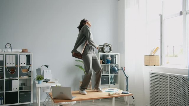 Slow motion of crazy girl dancing in office on desk taking off jacket throwing away wearing headphones. Work fun, crazy youth and lifestyle concept.