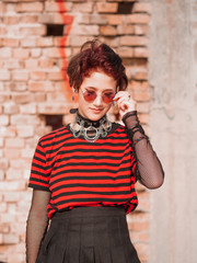 Emo girl on an brick background takes off hipster round glasses. Half cropped body