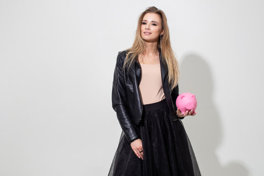 A girl in a black leather jacket holds a pink piggy bank on a white background