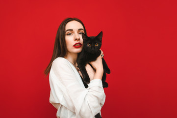 Fashionable portrait of a beautiful woman in a white shirt stands on a red background with a black cat in hand, looks into the camera with a serious face. Girl hugging a cat, isolated. Copyspace
