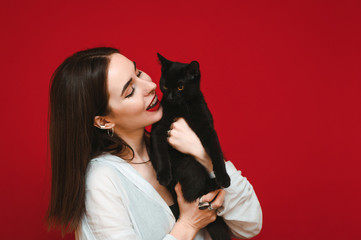 Closeup photo of positive owner with black cat on red background, playing with pet and smiling. Happy woman with cat in her arms isolated on red background. Copyspace