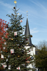 festlich dekorierter Weihnachtsbaum vor einem d&ouml;rflichen Kirchturm, blauer Himmer