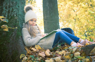 Cute little girl sitting with a book on her lap under the tree in public park. Girl sitting...