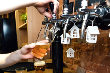 Man's hand pouring pint of beer behind the bar from the tap. Brick wall in the background. Toy houses of white color on the beer taps