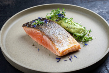 Minimalistic design salmon fish filet glazed with avocado and wasabi creme in a sliced cucumber as closeup on a modern design plate decorated with herbs