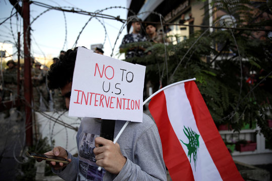A protester checks his mobile phone as he holds a sign and a Lebanese flag next to a barbed wire blocking the road to the U.S. Embassy in Awkar