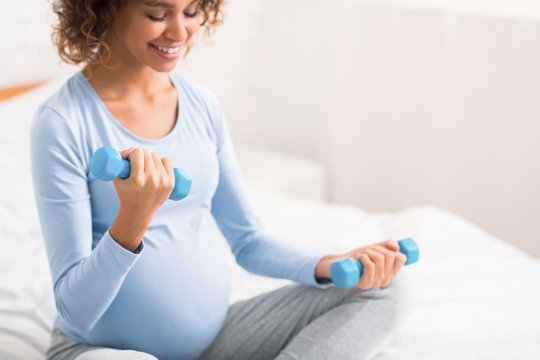 African pregnant lady exercising with dumbbells, sitting on bed