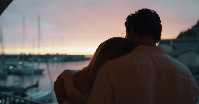 Close up of an young couple is enjoying time together on a terrace of a hotel room during their romantic vacation with panoramic view on a sea with yachts at sunset.