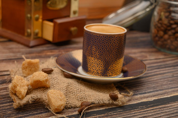 A Cup of coffee, pieces of brown sugar in a sugar bowl, coffee beans in a glass jar, a coffee grinder on a wooden background. Close up.