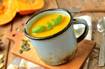 Old enamel cup of pumpkin soup with fresh basil and pumpkin seed on rustic wooden background