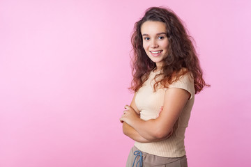 Portrait of cheerful confident teen girl with long curly brunette hair wearing casual style beige clothes standing with crossed hands and attractive smile. indoor studio shot isolated, pink background