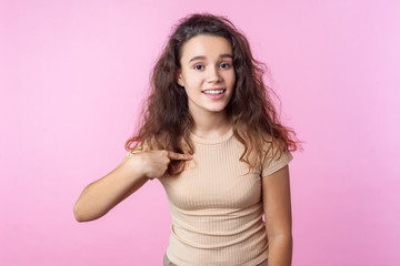 Portrait of lovely teenage girl with curly brunette hair wearing casual style beige clothes smiling at camera and pointing at herself, proud of achievements. studio shot isolated on pink background