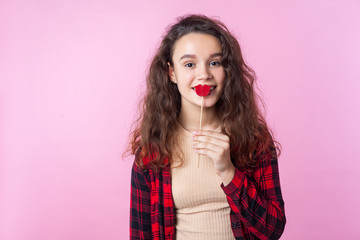Portrait of charming beautiful teenage girl with curly brunette hair in red plaid shirt smiling and covering mouth with red paper lips, valentines day. indoor studio shot isolated on pink background