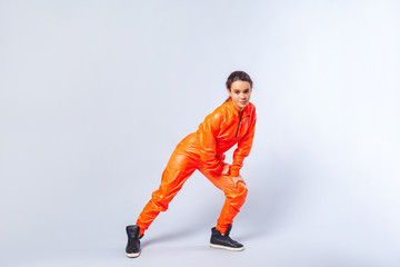 Fototapeta na wymiar Full length image of energetic teenage girl with brunette hair wearing bright orange jumpsuit dancing, showing butterfly hip hop move, hobby activities. indoor studio shot isolated on white background