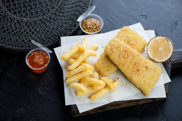 Breaded fish fillet with french fries on a wooden serving board, studio shot over black stone background
