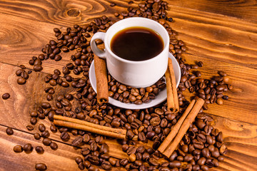 Cup of hot coffee, cinnamon sticks and scattered coffee beans on wooden table