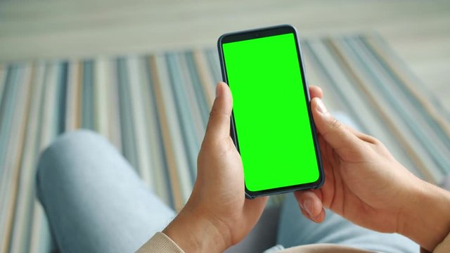 Male hands are holding contemporary smartphone with green chroma key screen sitting in apartment using device without touching screen. Modern technology and people concept.