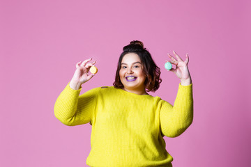 Cheerful plus size model posing with macaroon, fat girl loves sweets, pink studio background