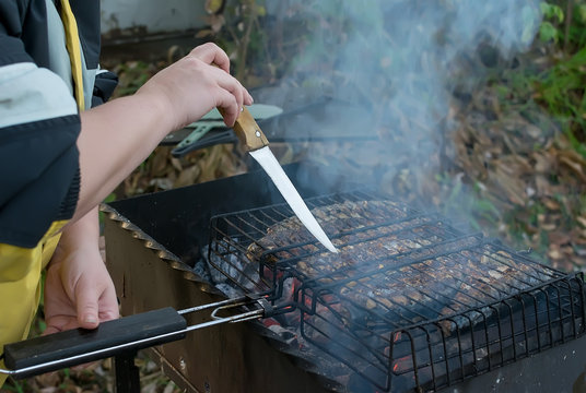 outdoors, the girl hand holds a knife and pokes it into the fish, which is fried on the grill barbecue on the grill